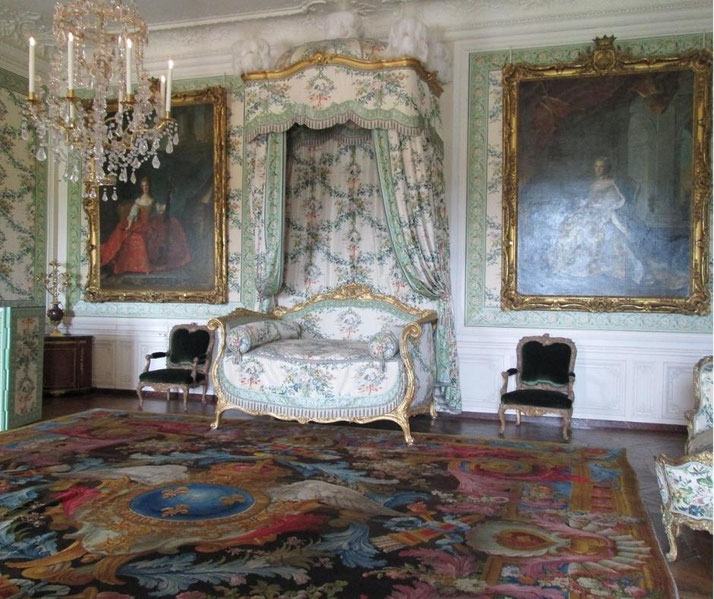 Elegant historic room with floral upholstered chaise lounge, framed portraits, chandelier, and richly patterned carpet, decorated in classical European style.