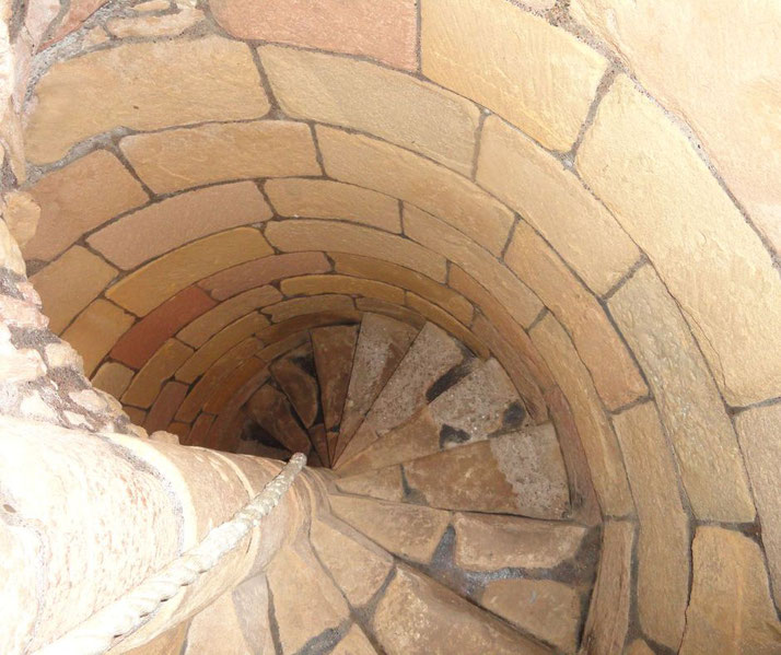 A spiral staircase made of stone steps, viewed from above. The walls are constructed with large, smooth stone blocks, and a rope railing can be seen along the edge of the stairs.