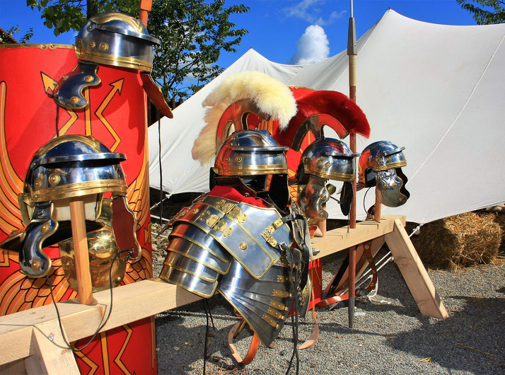 Display of Roman military armor and helmets on wooden stands with red shields and tents in the background.