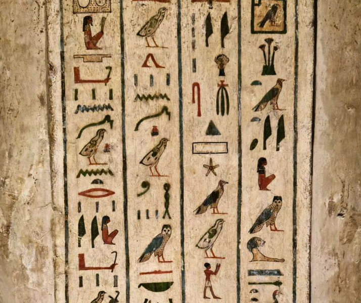 A colorful Ancient Egyptian hieroglyphic inscription in vertical columns, depicting birds, seated figures, plants, and abstract symbols on a weathered stone surface.