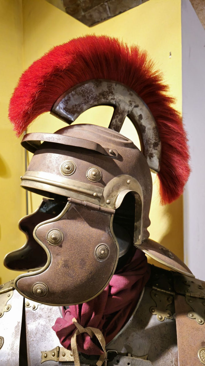 A Roman helmet with cheek guards, brass accents, and a red horsehair crest mounted on top, worn with segmented armor and a red scarf.