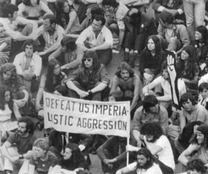 A group of protesters sit closely together holding a banner that reads “Defeat US Imperialistic Aggression” during an anti-war demonstration.