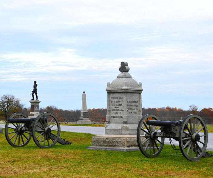 Civil War battlefield memorial with stone monuments, cannons, and a soldier statue on a grassy field under a cloudy sky.