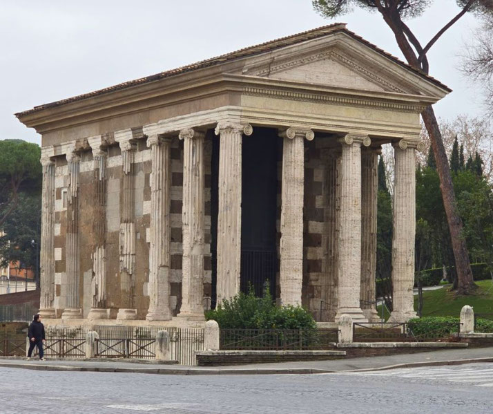 Well-preserved ancient Roman temple with Ionic columns, located in an urban park setting with trees and pavement.