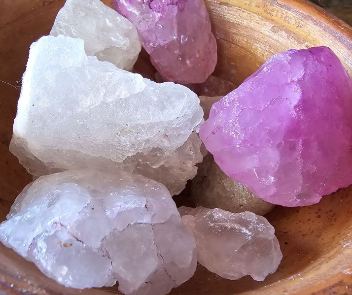 A wooden bowl filled with rough, translucent crystals, some clear or white and others vibrant purple. The stones have an unpolished texture, resembling quartz or calcite specimens.