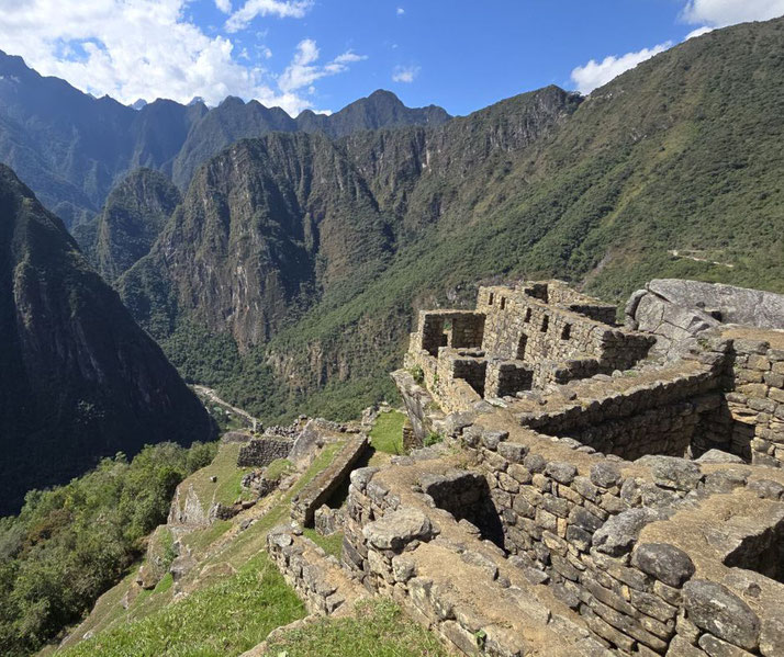 Ancient stone ruins of Machu Picchu sit atop a lush mountain ridge, overlooking a deep valley with towering green peaks under a bright blue sky. 