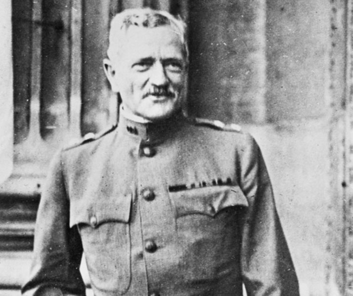 A military officer in uniform stands in front of a building, looking to the side with a slight smile, displaying a row of service ribbons on his chest.