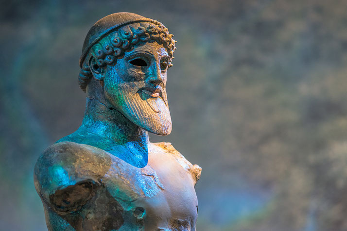 Close-up of an ancient Greek bronze statue with a bearded face, hollow eyes, and partial arms, lit with dramatic blue-toned lighting.
