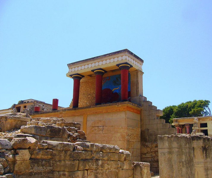 The ruins of the Palace of Knossos on Crete, featuring red columns, a reconstructed fresco, and stone walls.