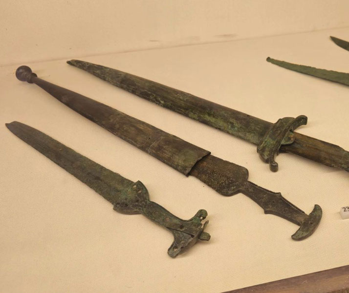 Ancient bronze daggers and swords with green patina, featuring ornate hilts and sheaths, displayed on a light surface.