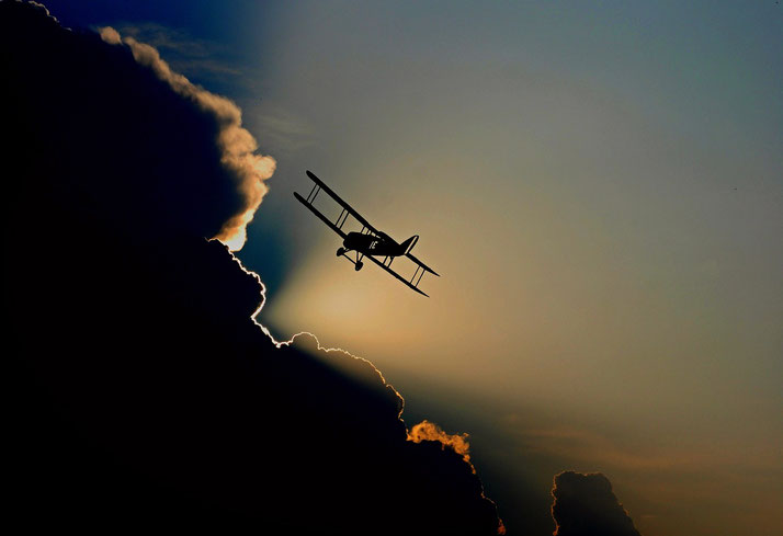 A biplane is silhouetted against the sky, flying near dramatic clouds with the sun glowing behind them.