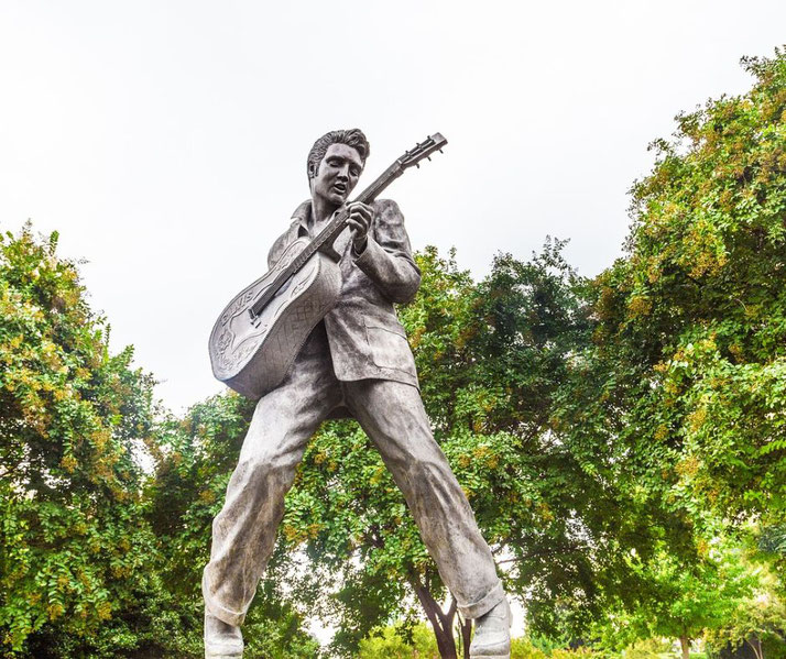 A large statue of a man playing guitar, striking a dynamic pose, stands among green trees against a cloudy sky.