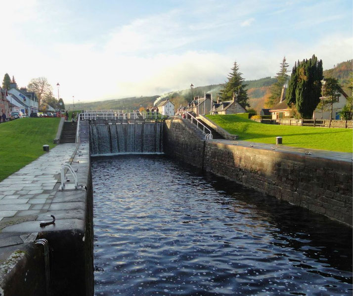 A scenic canal lock with cascading water, surrounded by lush green grass and quaint houses. The peaceful setting is enhanced by distant hills, lampposts, and a clear sky.
