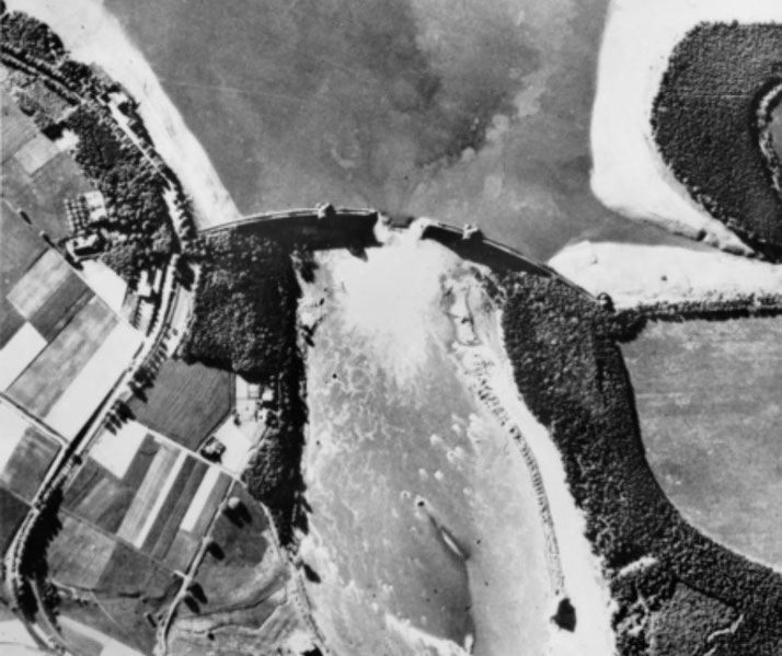 Aerial view of a breached dam with flooding downstream, showing surrounding farmland and landscape.