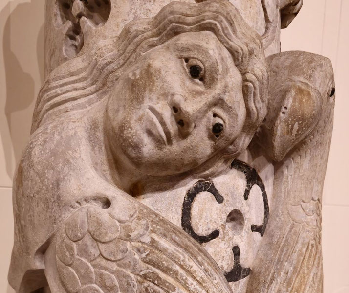 Weathered stone sculpture of a winged figure with long hair and wide eyes, holding a bird. A black circular design is carved on the chest.