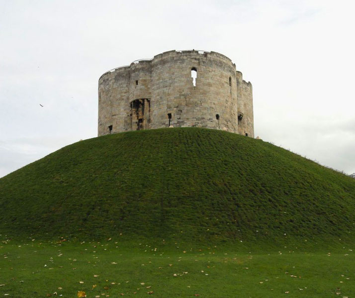 A circular stone tower on a large grassy mound. The tower has several small windows, and the surrounding landscape is green with scattered fallen leaves.