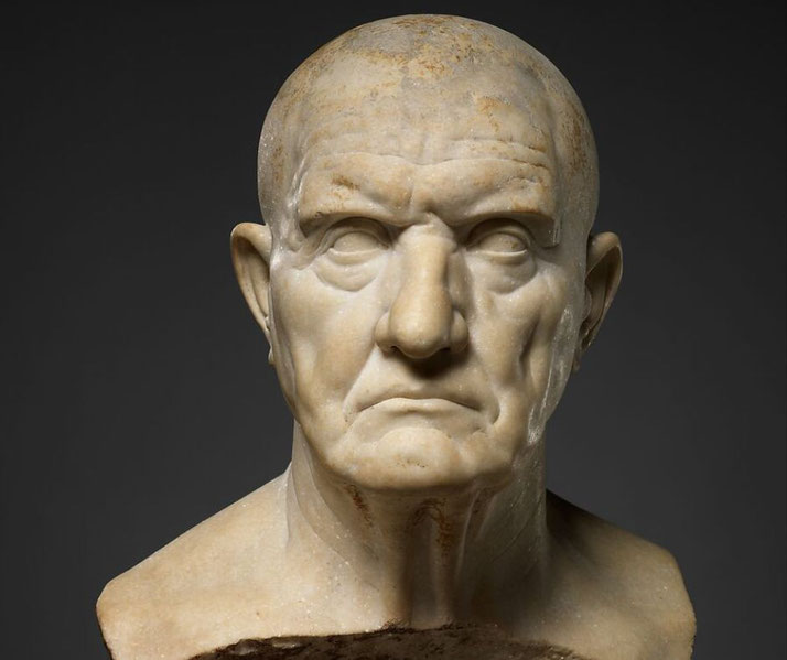 A marble bust of an older Roman man with a stern expression, deep wrinkles, and a bald head.