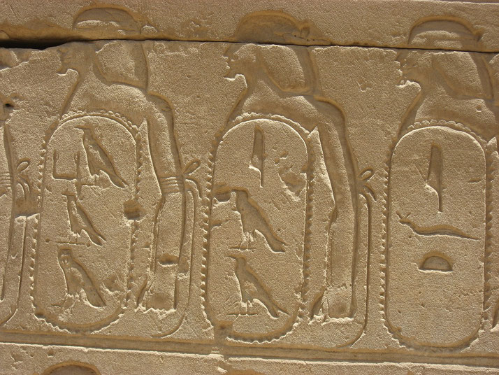 Stone wall with carved Egyptian cartouches containing hieroglyphs and figures, likely representing royal names or titles.