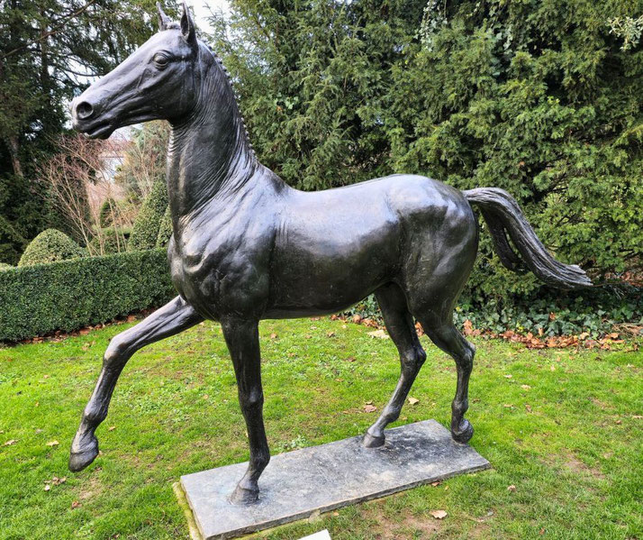 A bronze horse statue standing in a lush green garden. The horse is captured mid-stride, displaying elegant details and a lifelike stance, surrounded by neatly trimmed hedges and trees.
