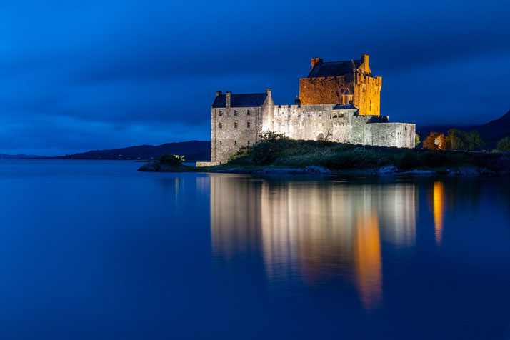 A stone castle lit up at night, reflecting on calm waters.