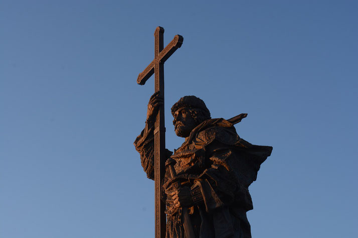 Bronze statue of a robed man holding a large cross, dramatically lit against a clear blue sky.