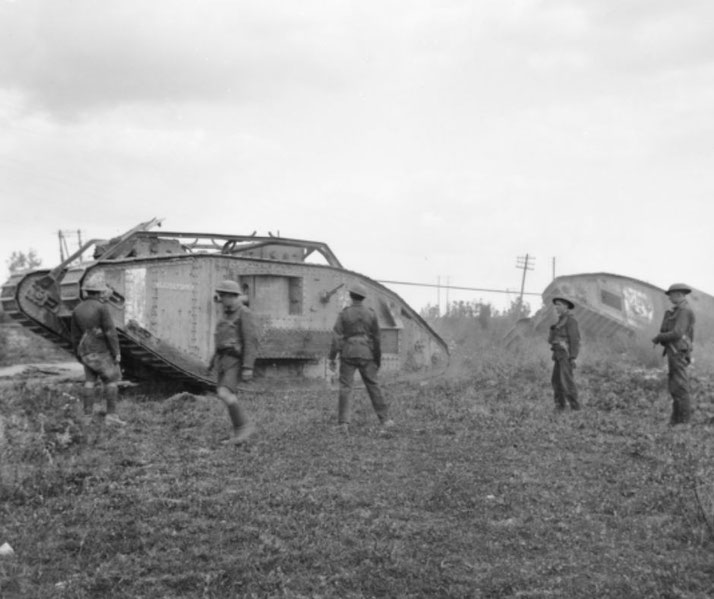 Black and white photo of soldiers standing near two WWI-era British Mark IV tanks on grassy terrain.