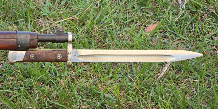 Close-up of a bayonet fixed to the end of a rifle, featuring a wooden grip and long, sharp blade, resting over green grass.