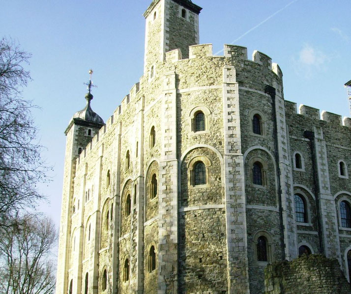 The iconic White Tower at the Tower of London, featuring its sturdy stone walls, arched windows, and battlements, standing tall under a bright blue sky with nearby bare trees framing the scene.