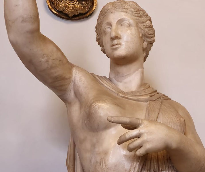 Marble statue of a woman with one arm raised and the other pointing forward, draped in a partially worn garment, showing classical Greek or Roman artistic style.