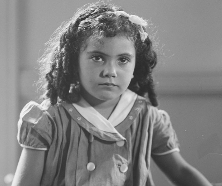A young girl with curly hair and a ribbon gazes seriously at the camera, dressed in a buttoned dress with a white collar.