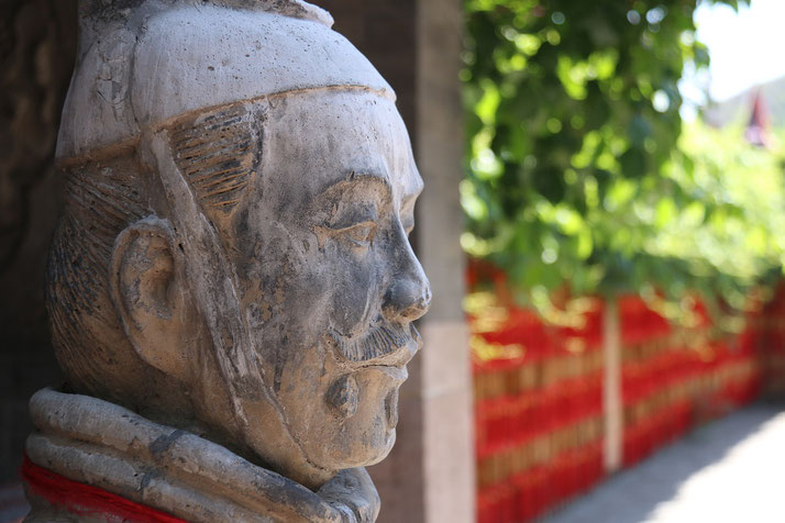 A close-up side profile of a weathered stone statue, possibly depicting an ancient warrior, with a blurred background of greenery and red accents creating a serene outdoor setting.