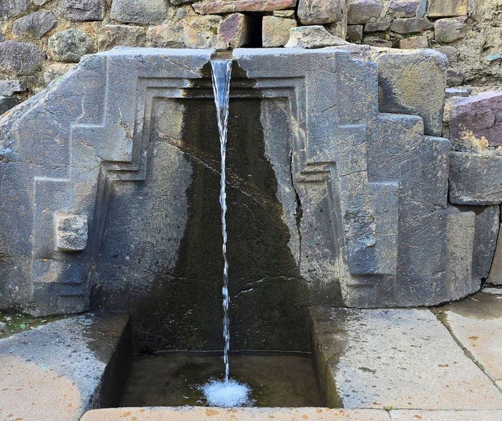 An ancient Incan water fountain, intricately carved from stone, with a symmetrical stepped design. Water flows smoothly from a narrow channel.