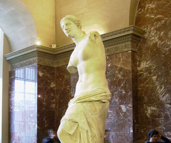 The Venus de Milo, a classical marble statue of a female figure with missing arms, stands in a museum setting.