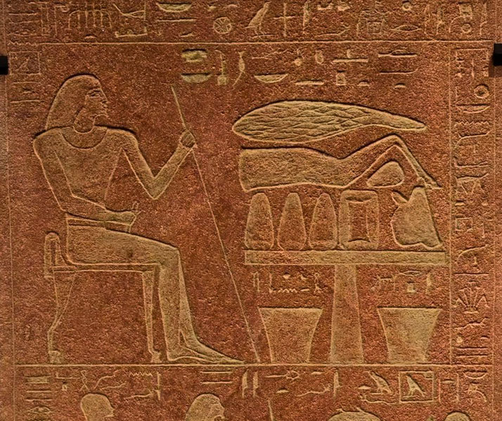 A carved Egyptian relief depicts a seated figure with a staff beside an offering table adorned with goods, possibly food or symbolic items. Hieroglyphs frame the scene, conveying its ritualistic significance.