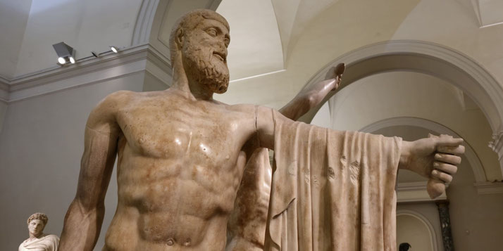 Large marble statue of a bearded man with muscular build, draped cloth over one arm, and arm extended forward holding an object, set in a classical hall.