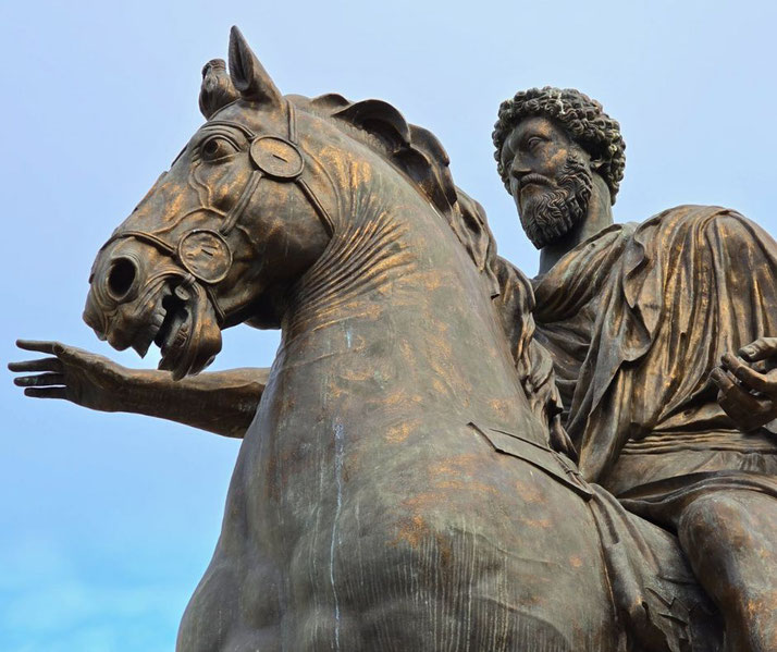 A close-up of a bronze equestrian statue depicting a bearded man in a flowing robe riding a horse. The horse's head is detailed with an open mouth and bridle, while the rider gestures with his right hand.