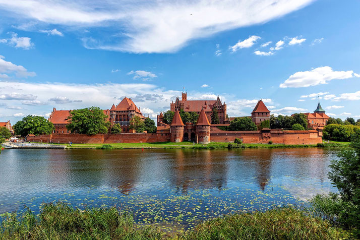 Large red-brick medieval castle complex with towers and walls reflected in a calm river under a bright blue sky.