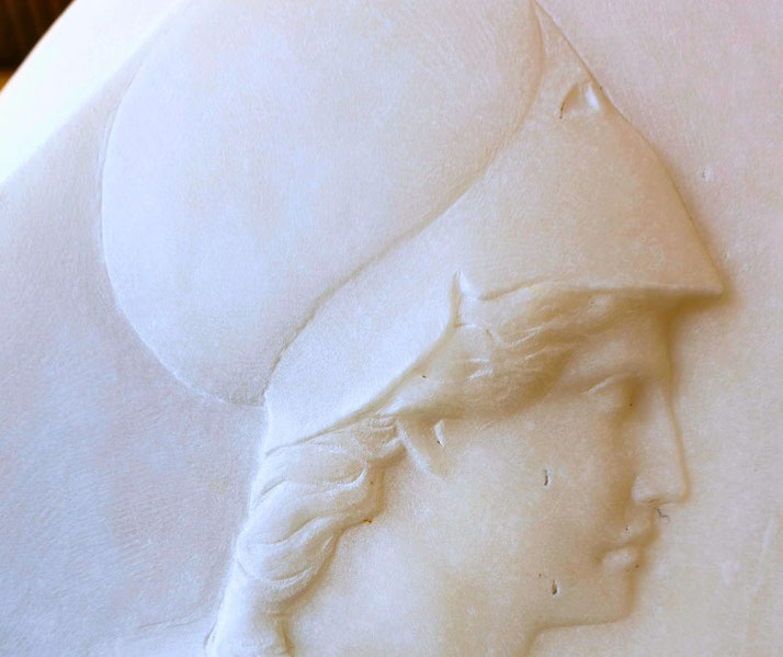 Relief carving of a classical figure in profile wearing a crested helmet, likely representing a warrior or deity, on smooth marble.