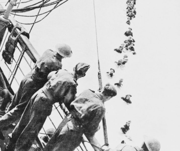 Soldiers aboard a ship watch others wading through water in a long line during a World War II evacuation.