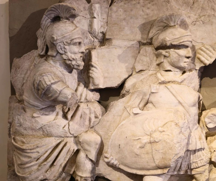 Fragmented marble relief of two Roman soldiers in armor and crested helmets, one holding a shield with a lion motif, captured mid-action in a battle scene.