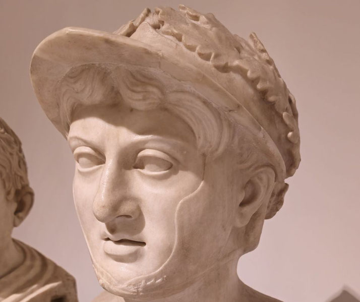 Marble bust of a young man with soft features, wearing a laurel-crowned helmet; delicate facial details and short wavy hair.