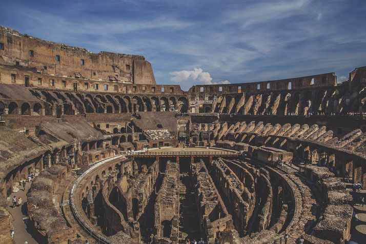 The iconic Roman Colosseum stands under a bright blue sky, revealing its ancient, tiered architecture and central arena with visible underground passages, bustling with visitors exploring its history.