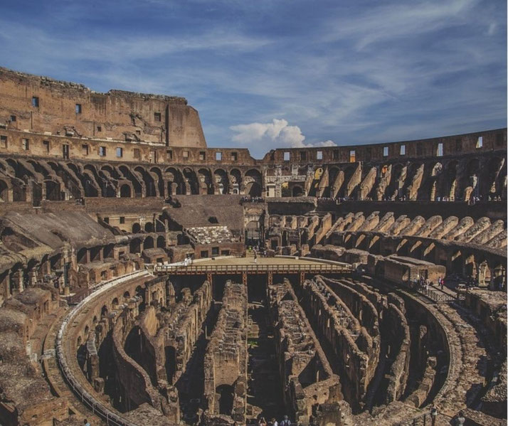 The iconic Roman Colosseum stands under a bright blue sky, revealing its ancient, tiered architecture and central arena with visible underground passages, bustling with visitors exploring its history.