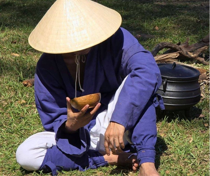 A person in traditional clothing wearing a straw hat, sitting on the grass while drinking from a wooden bowl. A black pot and firewood are nearby, suggesting an outdoor cooking scene.