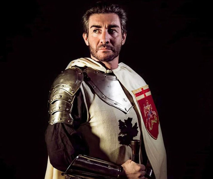 A man dressed as a medieval knight wears a white cape with a red heraldic emblem, metal shoulder armor, and a black tunic.