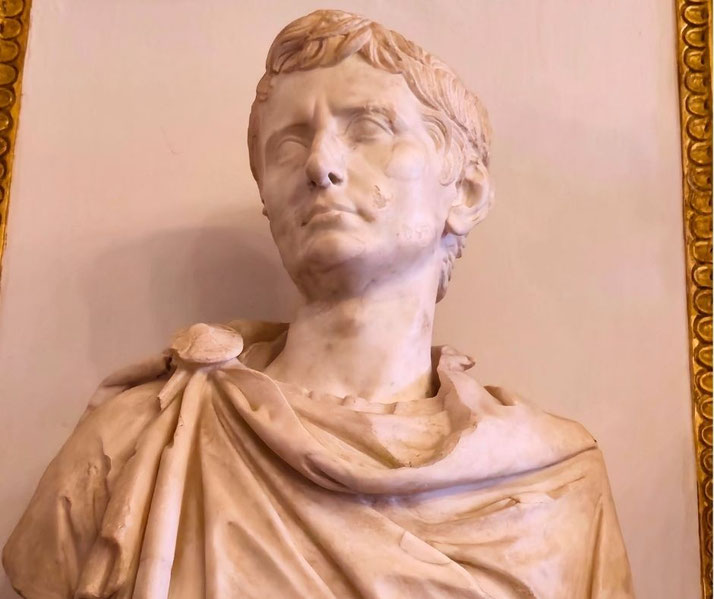 A marble bust of a Roman man with short hair and draped toga, displayed indoors against a plain wall with a decorative frame edge.