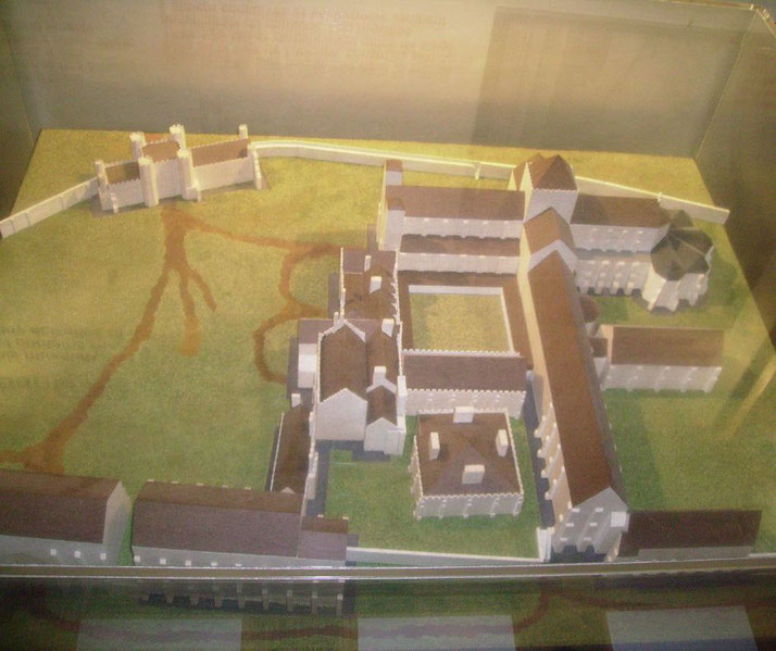 A scaled-down model of a medieval abbey complex, featuring detailed walls, towers, courtyards, and surrounding green terrain, displayed under protective glass.