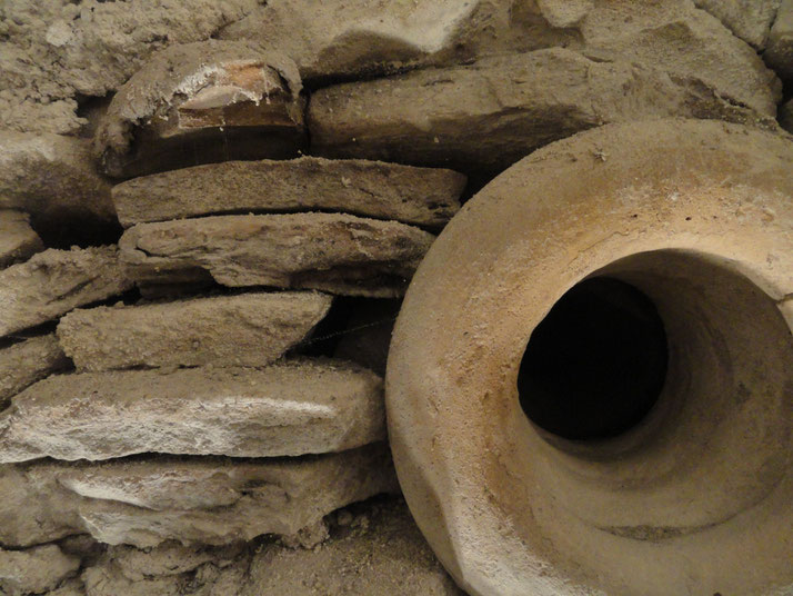 Close-up of ancient stacked stonework beside the mouth of a large clay vessel, both covered in dust and partially embedded in soil.