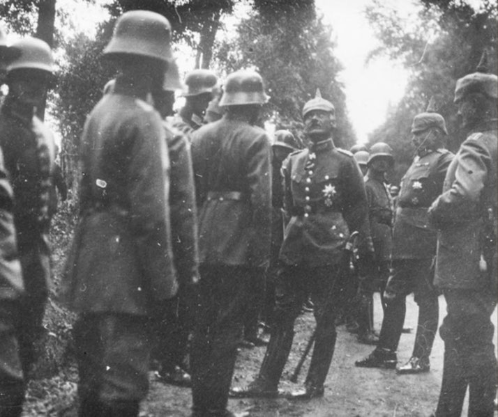 A group of German soldiers in uniform and helmets stand in formation on a wooded path, with a decorated officer inspecting or addressing them.