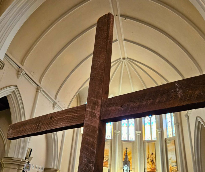 A large wooden cross inside a church, set against a vaulted ceiling and stained-glass windows.